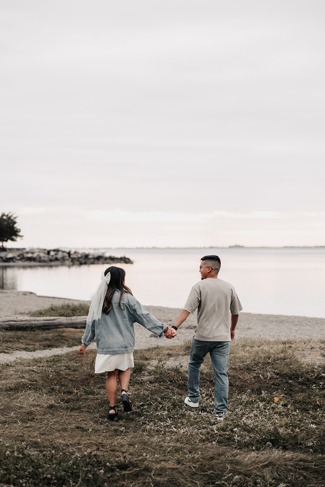 The couple walks away from the camera, hand-in-hand, towards the calm waters of the beach. The woman wears a veil, and their relaxed attire adds a casual yet romantic touch to this beach engagement photo.