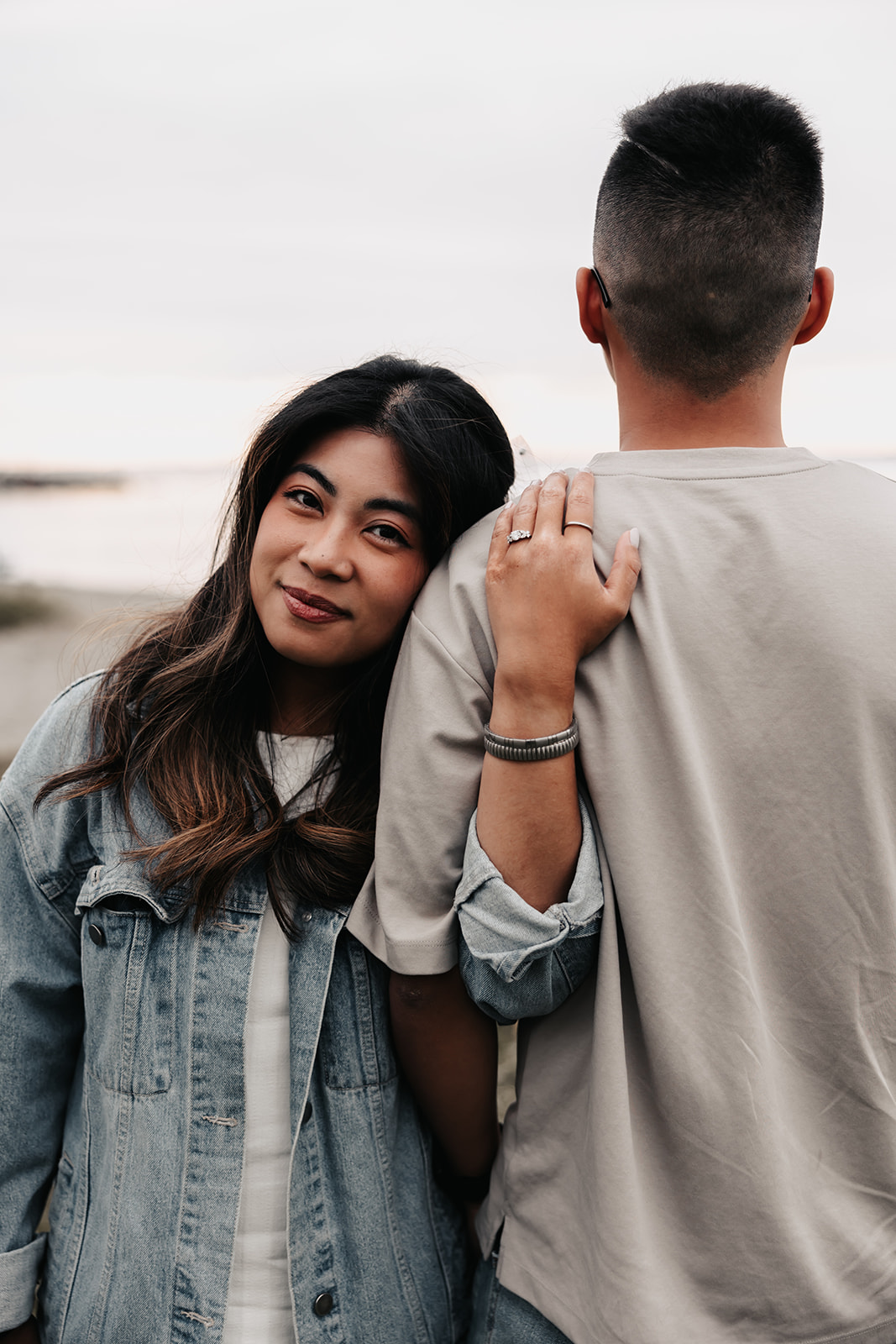 The woman leans gently on her partner's back, her hand resting on his shoulder and showing off her engagement ring. She looks at the camera with a soft smile during this intimate beach engagement moment.