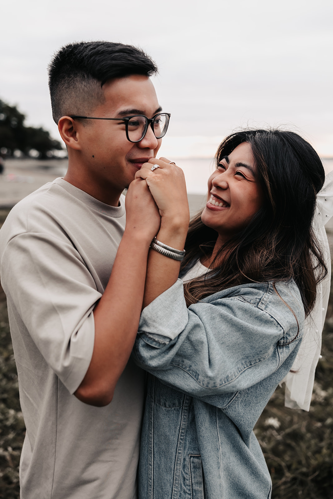 The couple shares a light-hearted moment, with the man holding the woman's hands while they laugh together. Their casual, candid expressions capture the joy of their beach engagement photos.