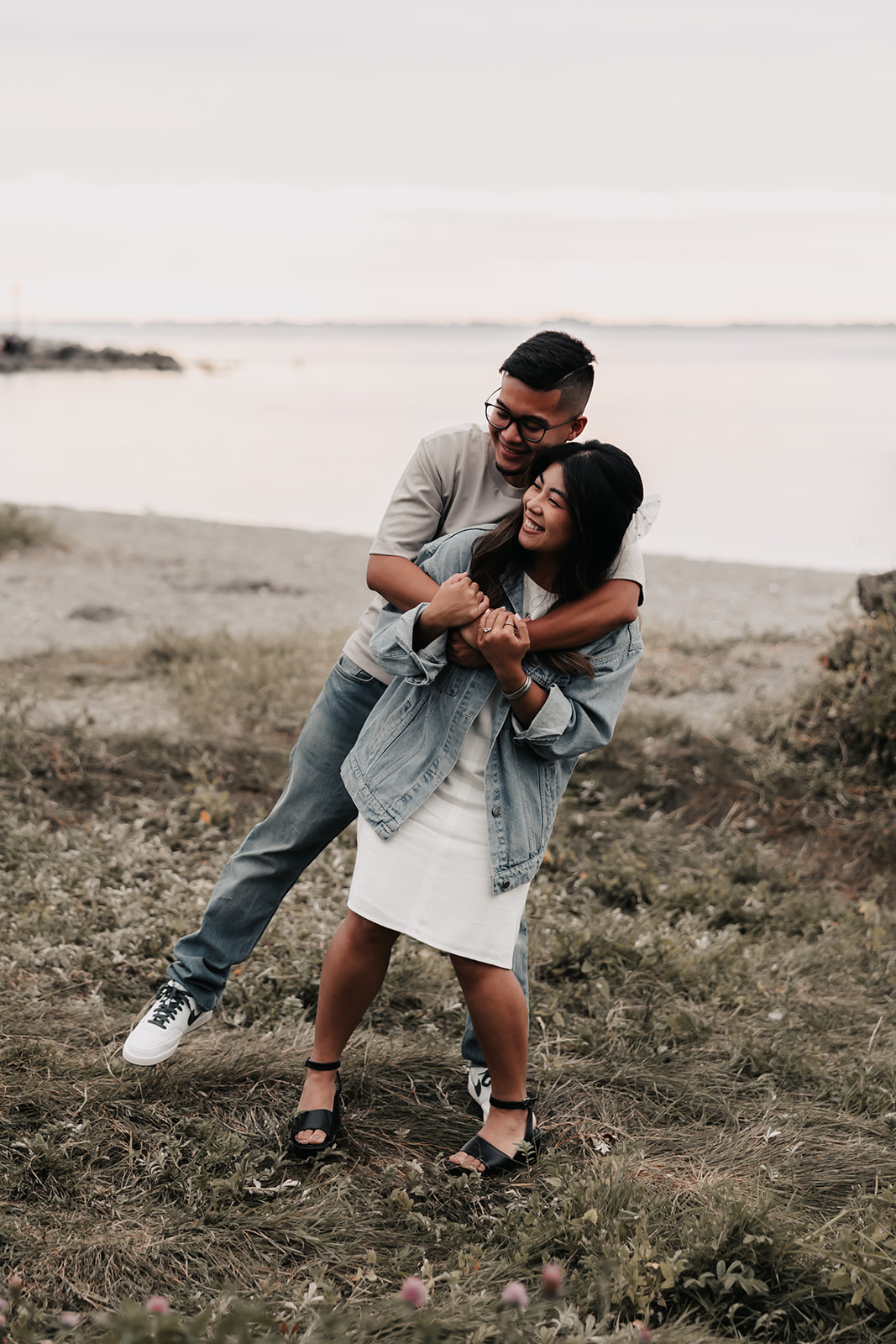 The man wraps his arms around the woman from behind as they stand on the beach, both smiling and laughing. Their playful energy shines through this beach engagement photo.