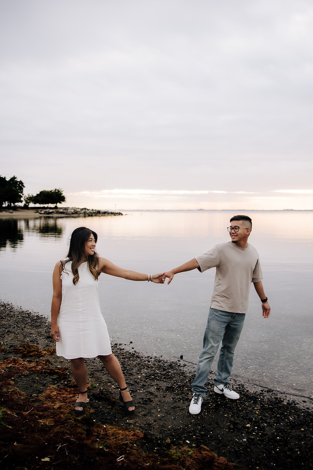 The couple holds hands, with the woman playfully pulling the man forward along the shoreline. The ocean serves as a peaceful backdrop for this playful beach engagement photo.