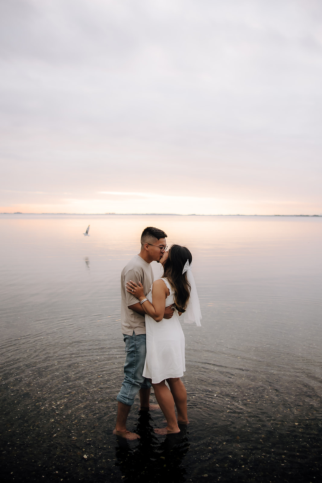 A couple embraces in the water during sunset, with the woman sitting on the man's lap. The soft colors of the sunset reflect on the calm water, creating a serene backdrop for their intimate moment.