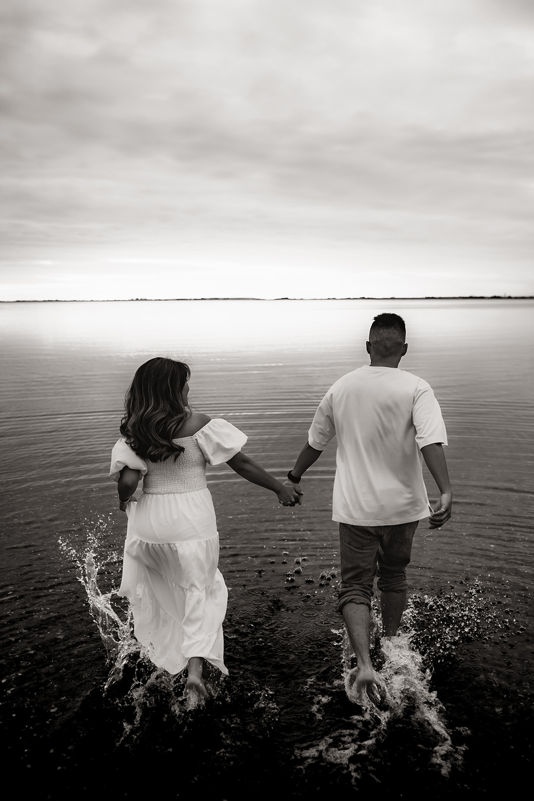 The couple holds hands, running into the water, captured in black and white during their adventurous beach engagement session.