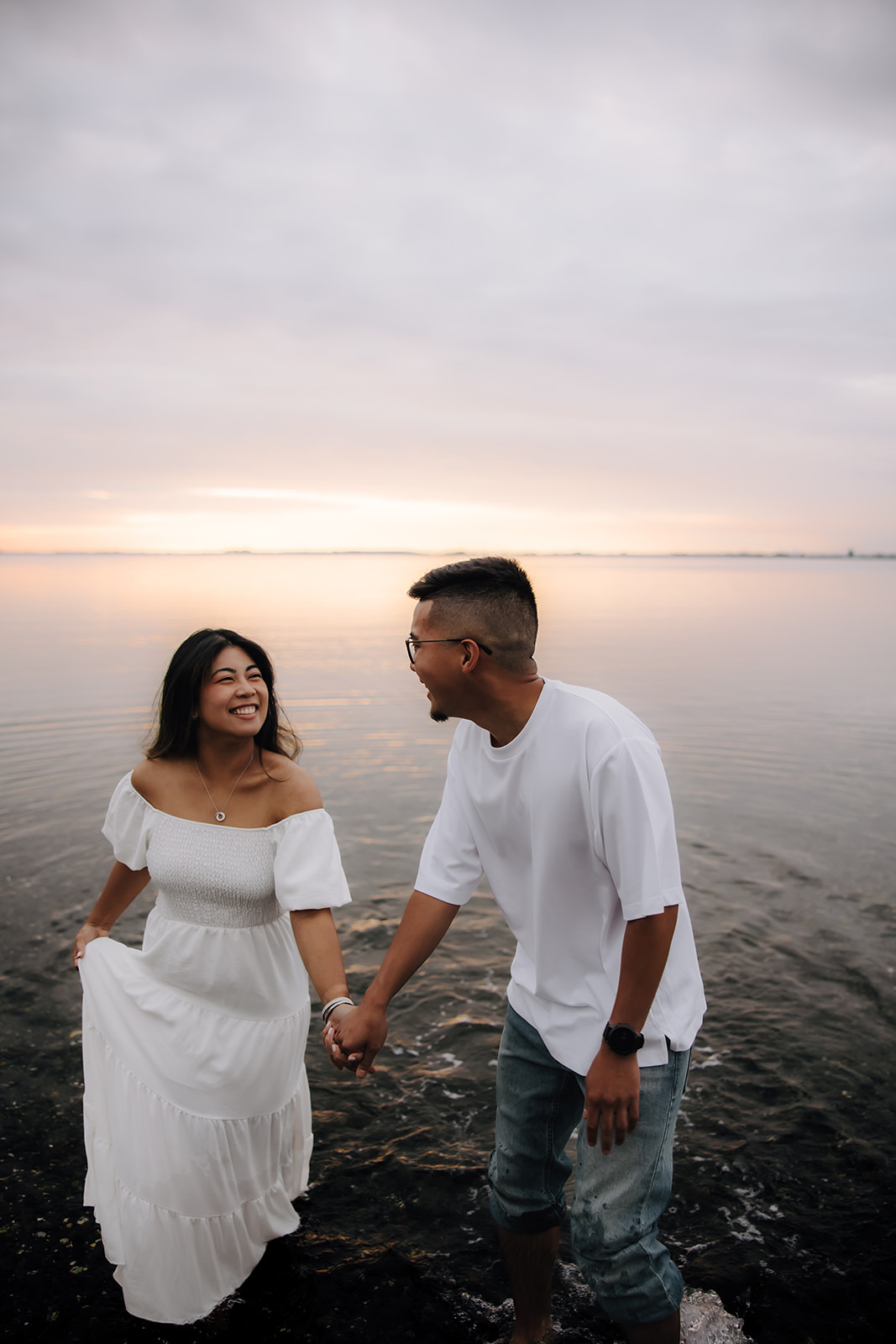 A joyful couple walks through the shallow water, smiling and laughing together. The sunset casts a warm glow on their faces, highlighting their happiness during this beach engagement photo session.