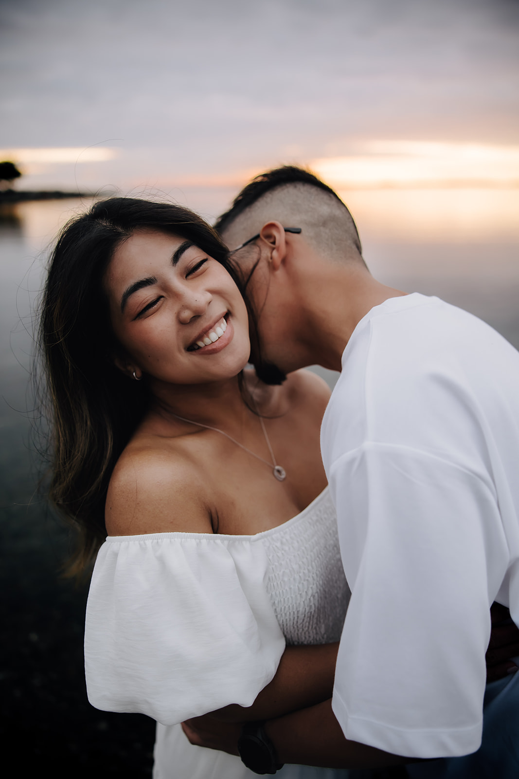 Groom to be kissing bride to bes neck as she smiles as they stand in the ocean