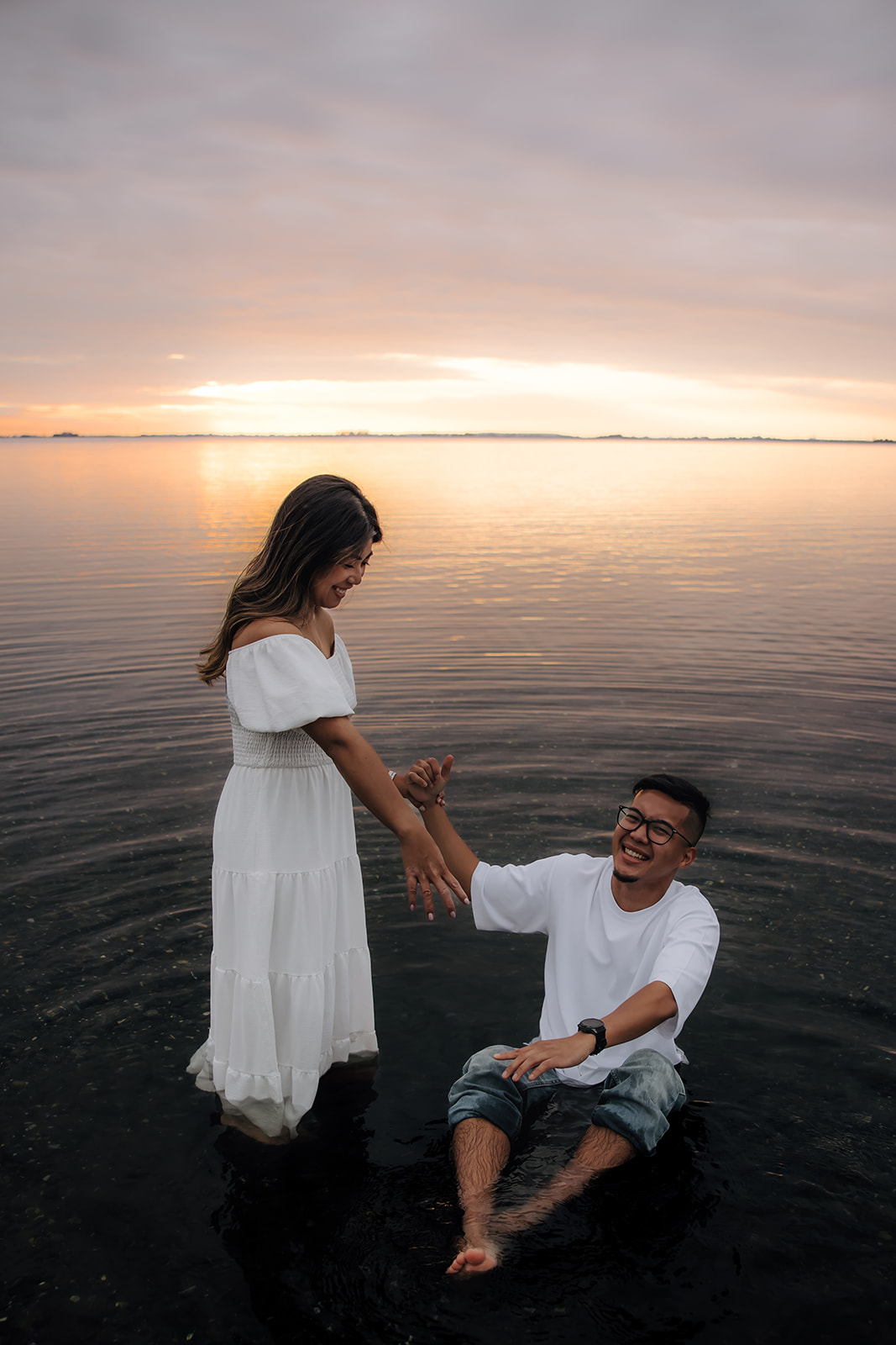 The bride-to-be helps her groom-to-be out of the water, laughing together during a playful moment in their beach engagement photos.