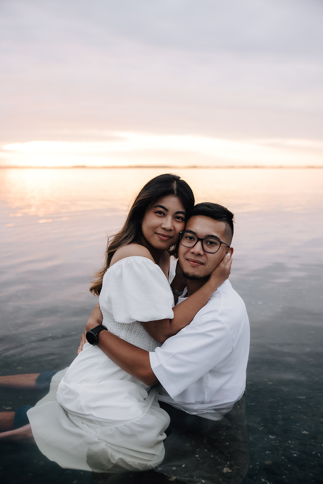 A couple sits in the water, holding each other as they smile softly toward the camera, capturing a quiet, intimate moment during their beach engagement photo session.