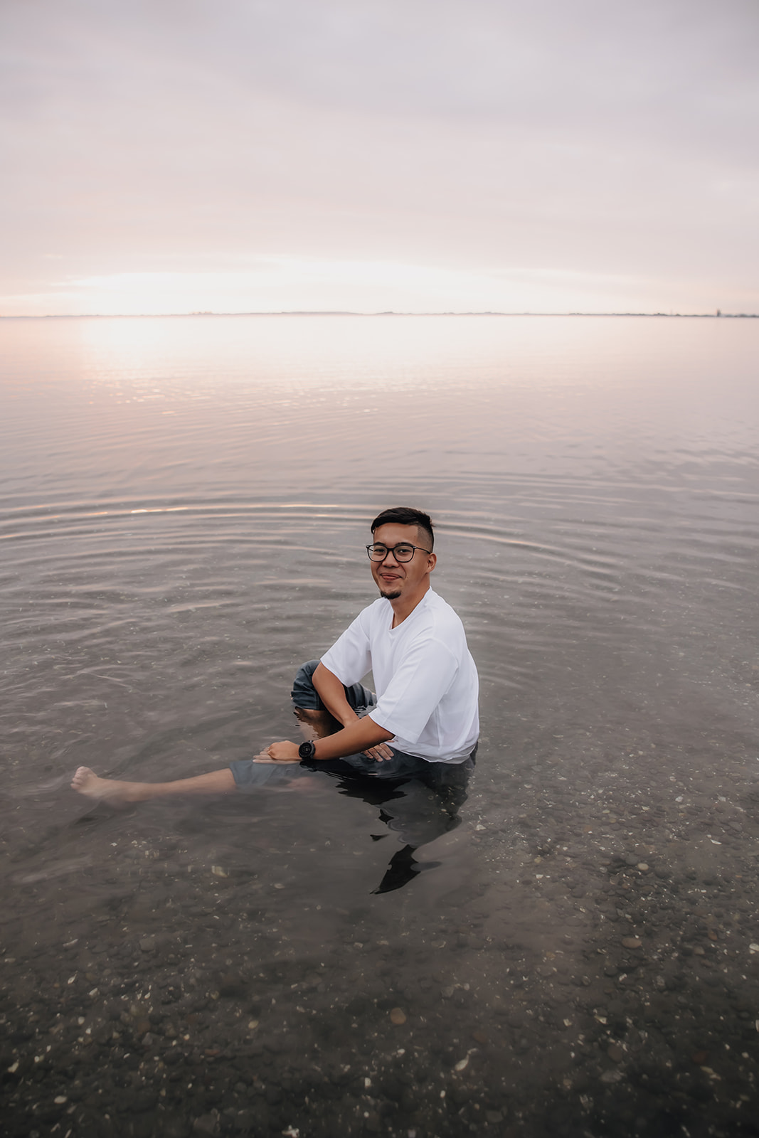 A man sitting in shallow water during a beach engagement photo session, smiling at the camera as the calm water reflects the soft sunset in the background.