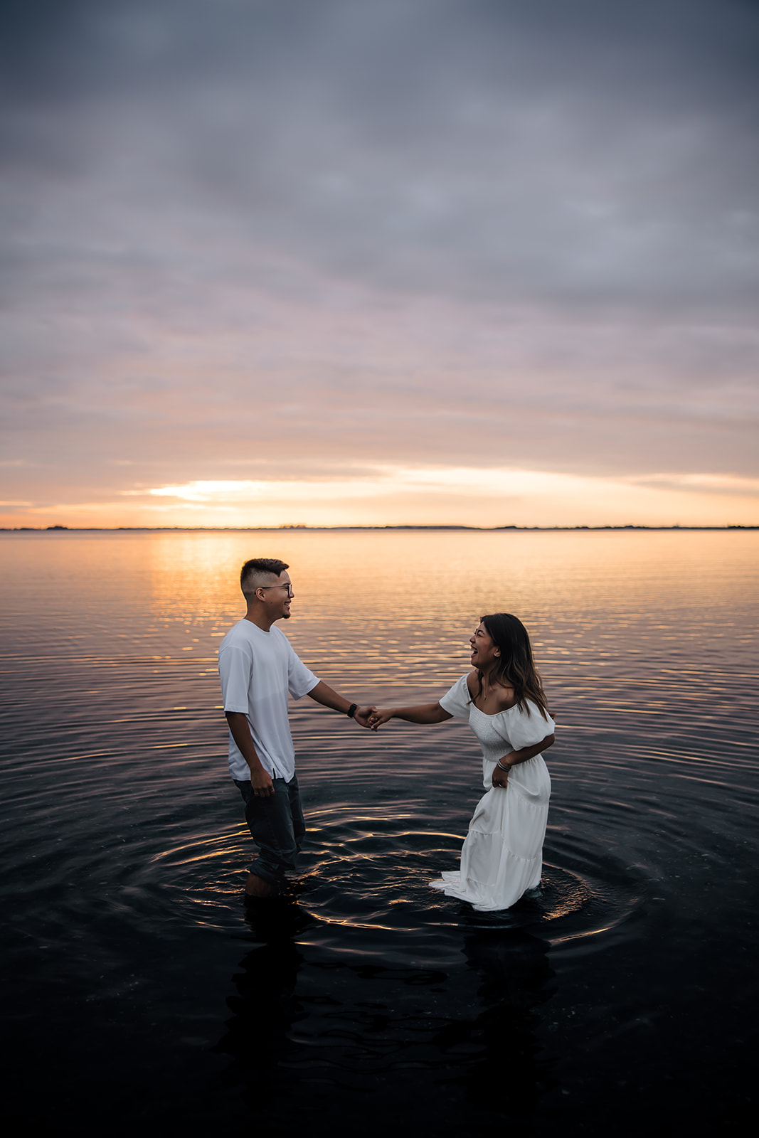 A couple holding hands in knee-deep water during a beach engagement photo at sunset, laughing and sharing a candid moment in the peaceful water.