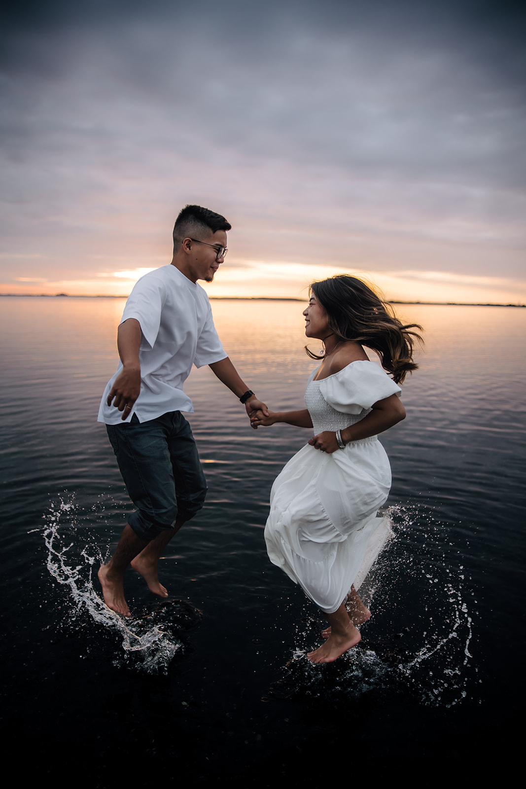 A couple playfully running and jumping in the water at sunset during a beach engagement photo session, creating splashes and smiles under the evening sky.