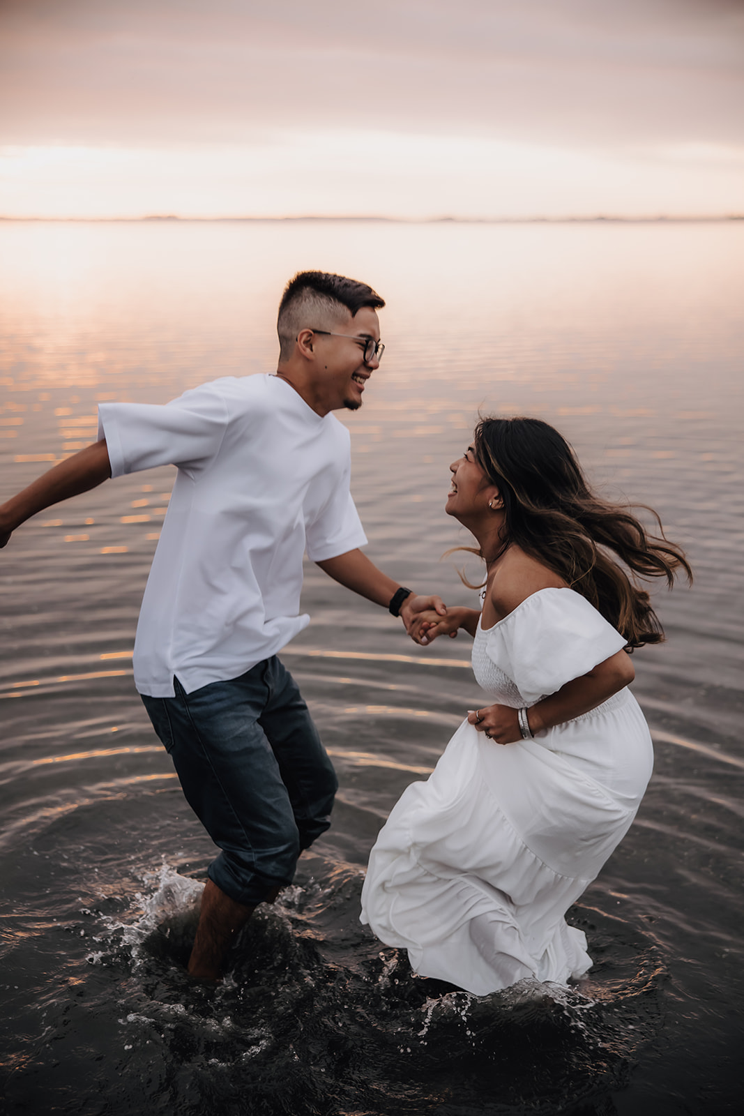 A man and woman holding hands and playfully splashing in the water during a fun and candid beach engagement photo, showing their joyful energy as the sun sets behind them.