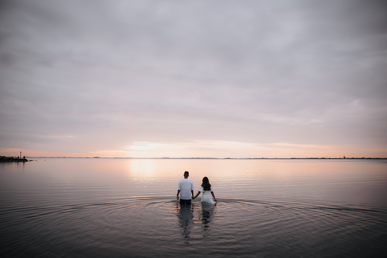 The couple walks hand in hand into the ocean, captured from behind, creating a serene and intimate beach engagement photo with a soft pink and blue sky at sunset.