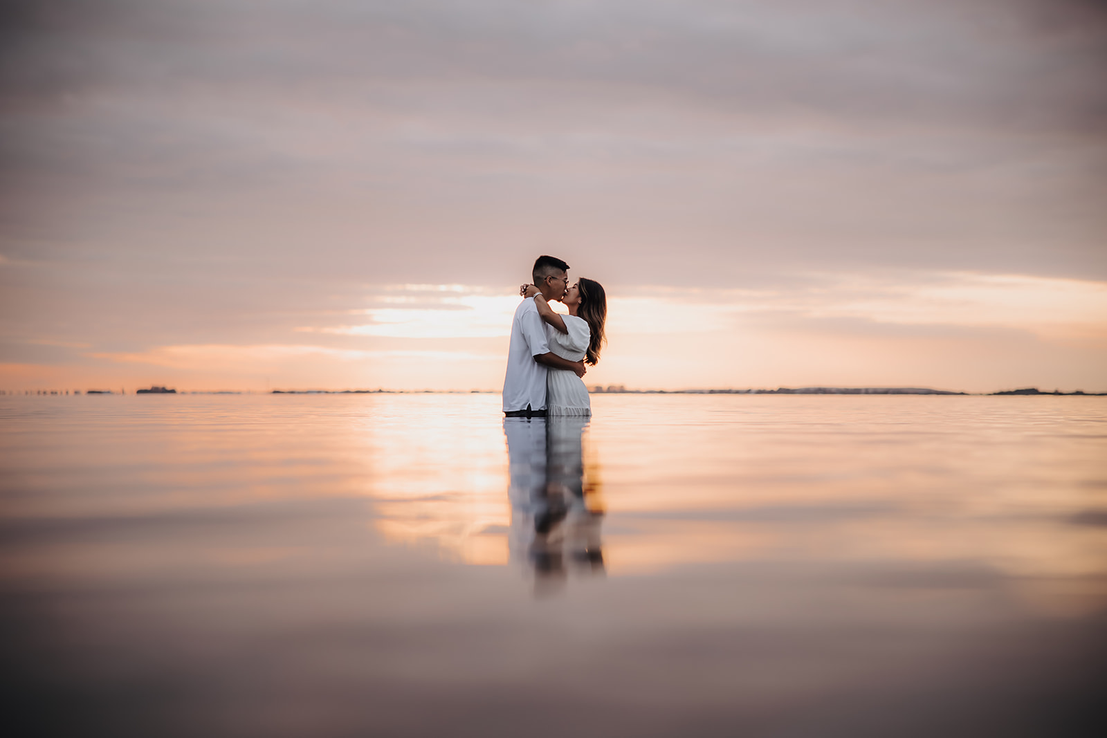 A romantic beach engagement photo of a couple embracing in knee-deep water at sunset, with a reflection of soft pink and golden hues on the water.