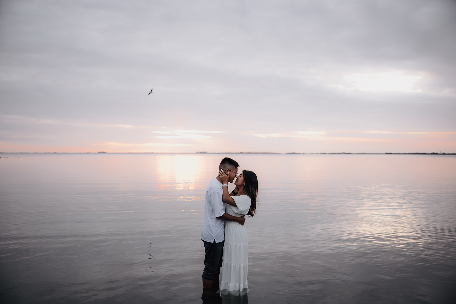 The couple shares an intimate kiss while standing in the water, with the vast ocean and sky blending together in soft tones, creating a magical sunset beach engagement moment.