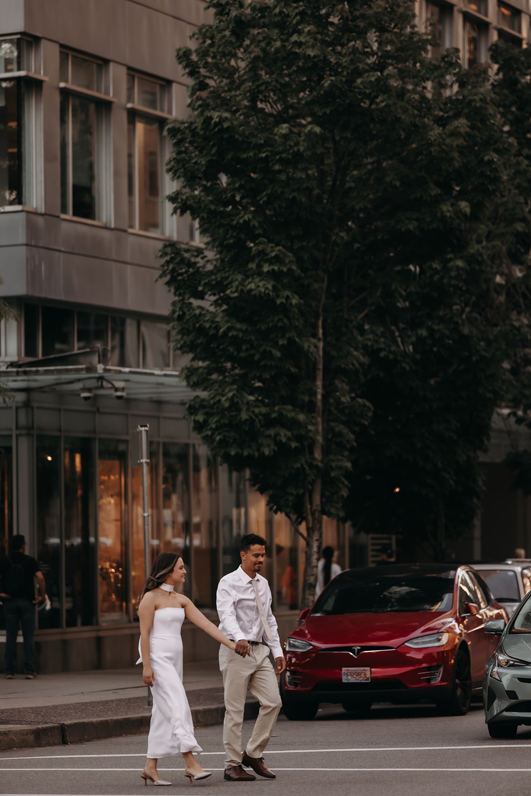 A couple walking hand-in-hand across the street, surrounded by modern buildings in a downtown engagement photo.