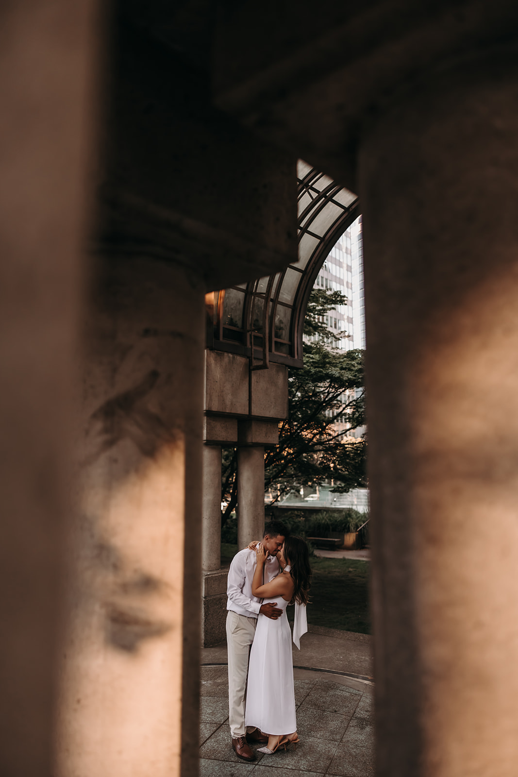 A couple standing between concrete columns, sharing an intimate embrace in a downtown setting.