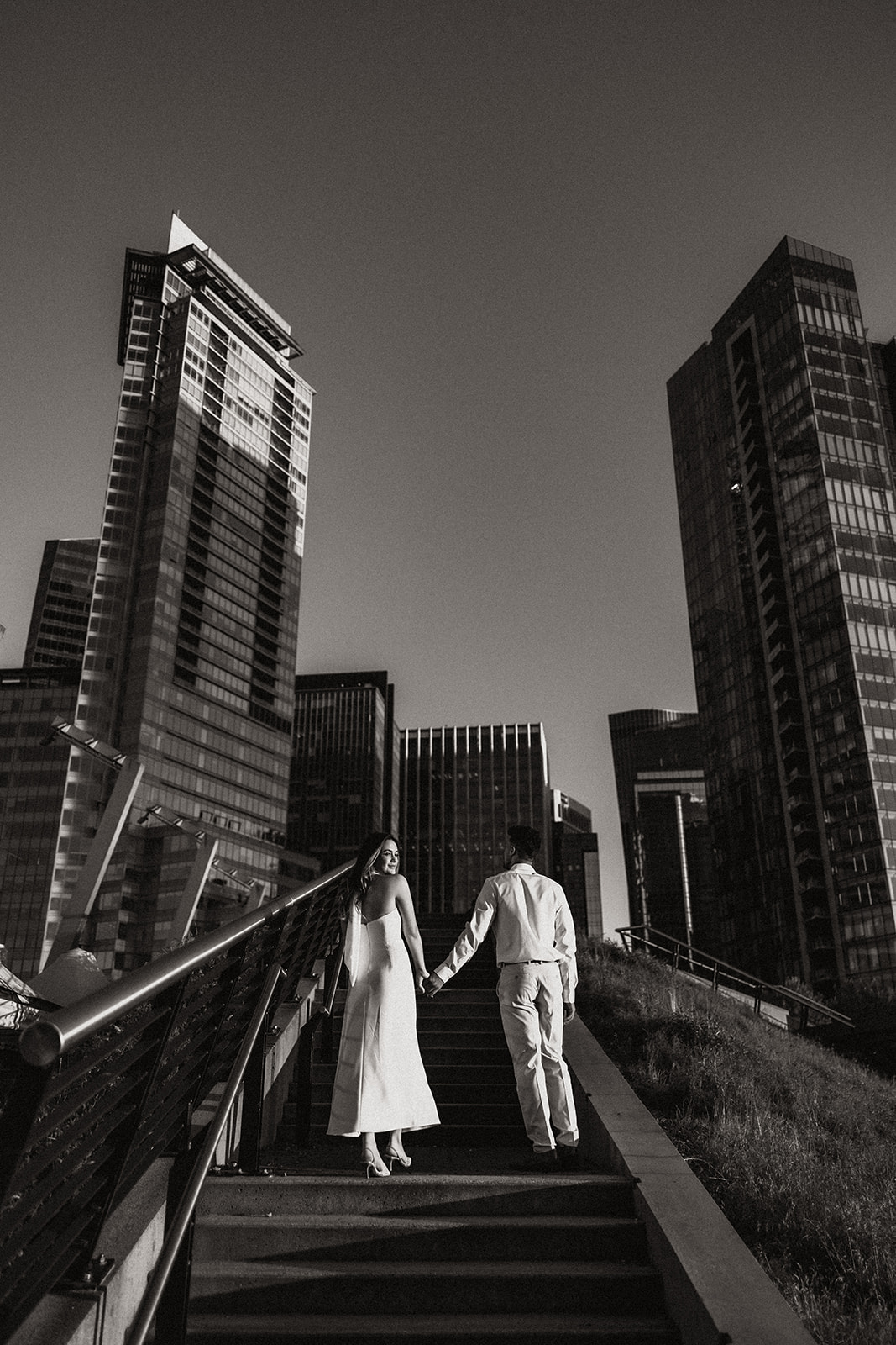 A couple holding hands, walking up the stairs with tall skyscrapers in the background.