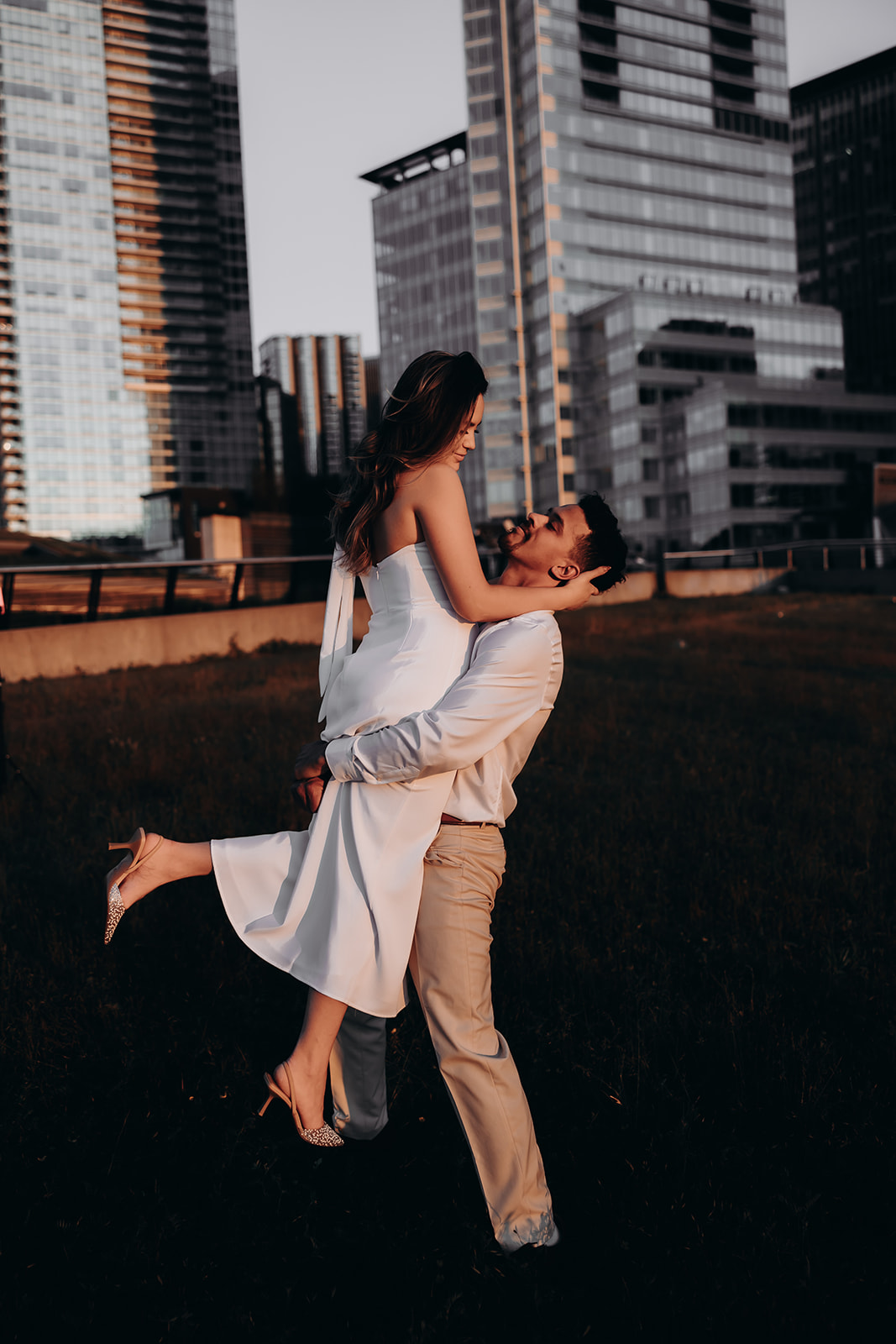 The groom-to-be holds his bride in the air as they share a candid moment against the backdrop of city skyscrapers during their downtown engagement photoshoot.