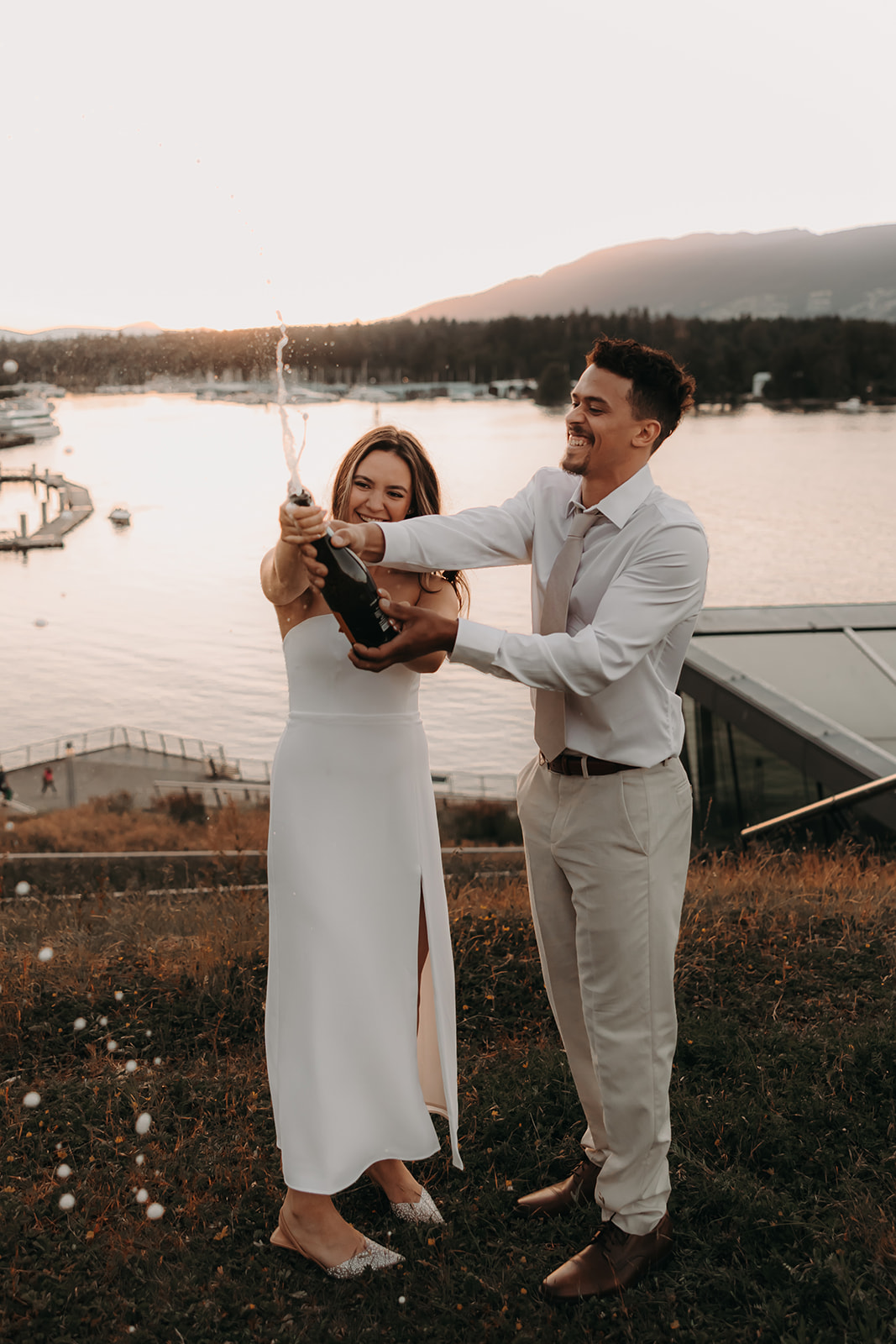 The couple laughs together while spraying champagne during their engagement photoshoot, with the city skyline and waterfront in the background.