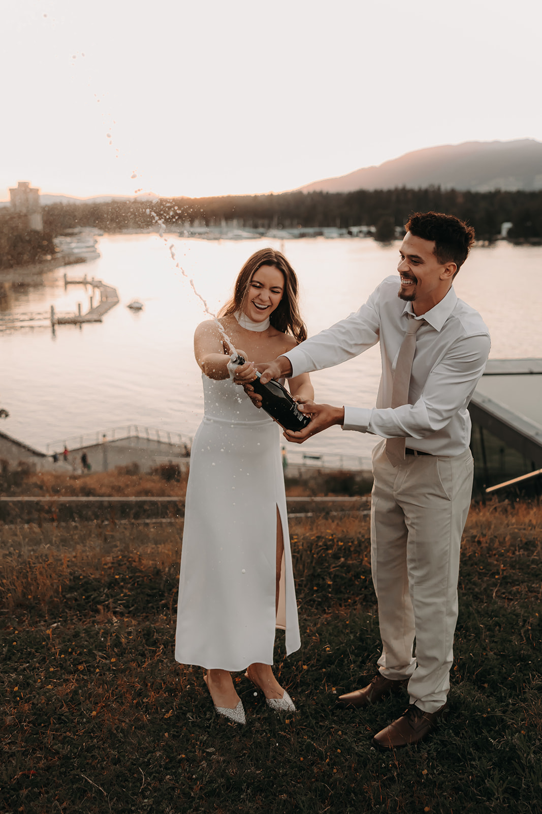 The couple joyfully pops champagne during their engagement photoshoot by the water, celebrating their love against the backdrop of a downtown cityscape.