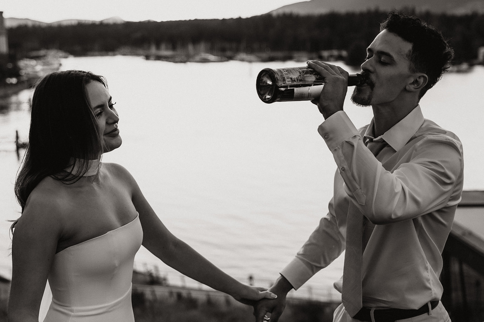 A groom takes a swig of champagne as the bride smiles, sharing a playful moment by the water during their downtown engagement session.