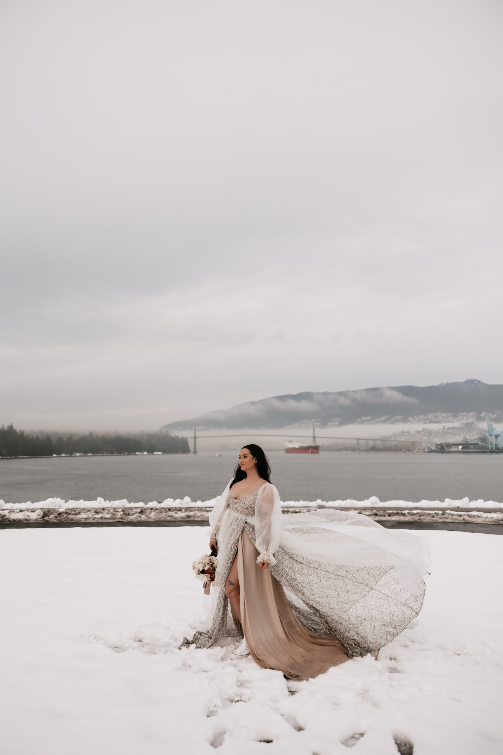 The bride stands on a snowy shoreline, her gown flowing in the wind with the Vancouver harbor in the background.