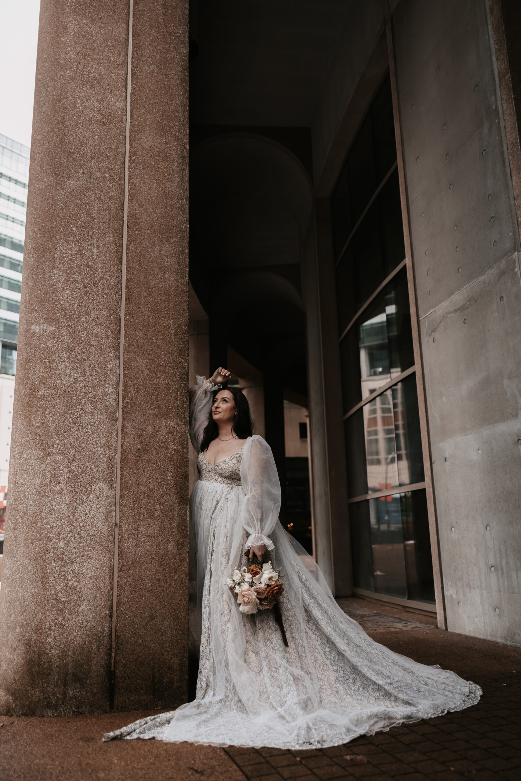 A bride leaning against a tall stone column in her elegant wedding gown, holding a bouquet of roses.