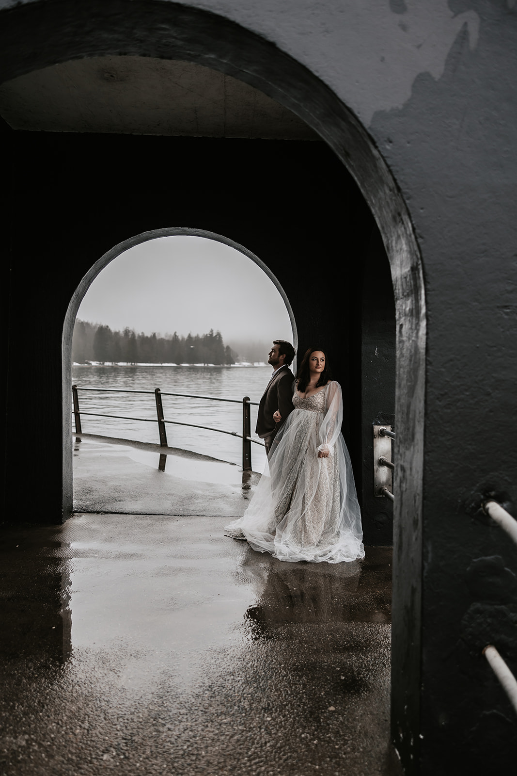 A couple stands in front of an arched structure near the water on a foggy day, capturing the serene mood of their Vancouver elopement.