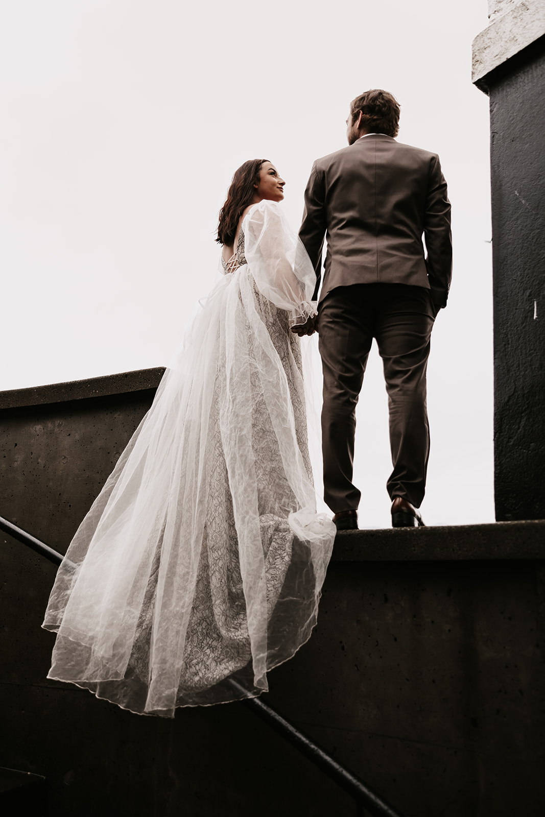 The couple holds hands as they prepare to descend the stairs, their elegant attire making a statement against the urban backdrop.