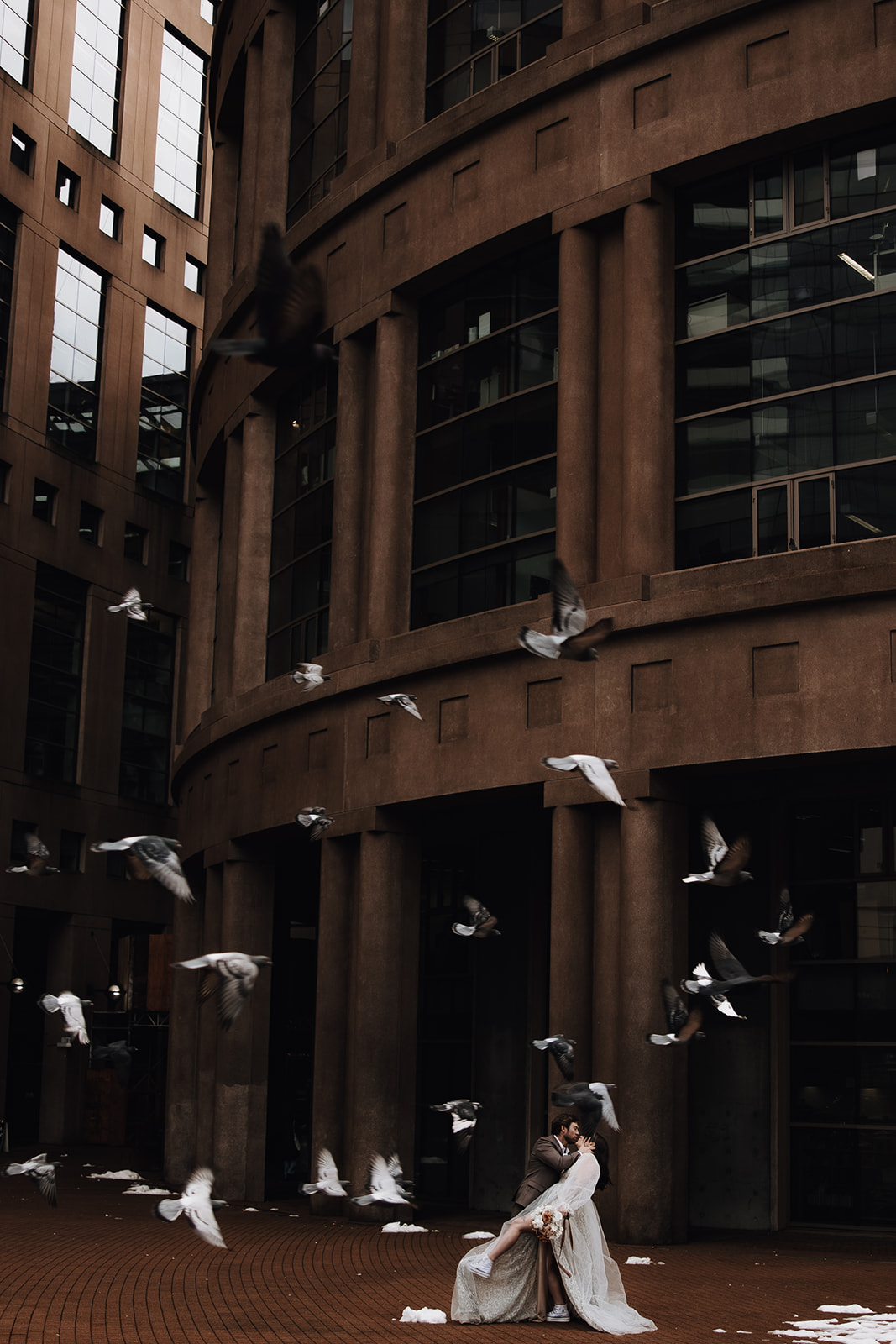 Birds flying around the couple embracing outside a modern building, capturing a candid Vancouver elopement moment.