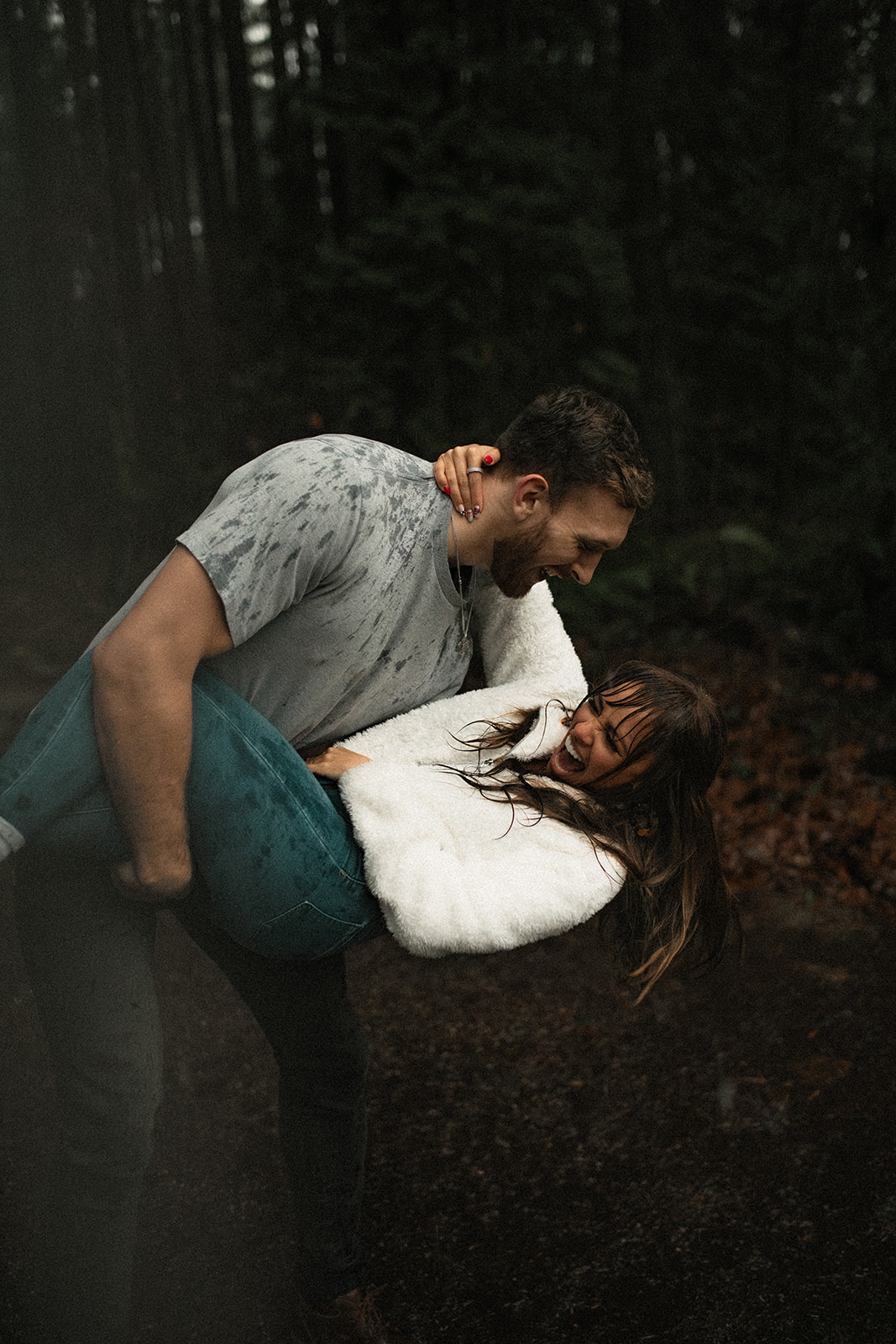 A couple laughing and enjoying each other's company in a rainy day couple picture. The woman is playfully lifted off the ground by her partner, showcasing their fun couple photoshoot ideas amid the lush greenery of the Pacific Northwest.