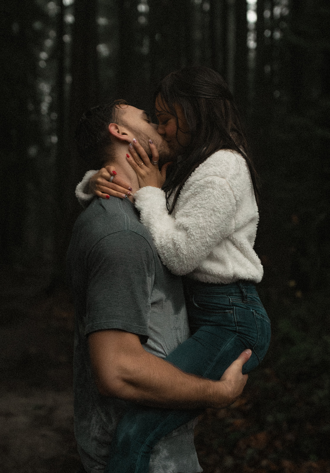 A couple shares a passionate kiss in a rain-soaked forest, embodying the essence of rainy day couple pictures. The man holds the woman securely in his arms, showcasing their playful connection. Her fluffy white sweater contrasts beautifully with the dark, moody backdrop of tall trees, creating a stunning visual that highlights this fun couple photoshoot idea.