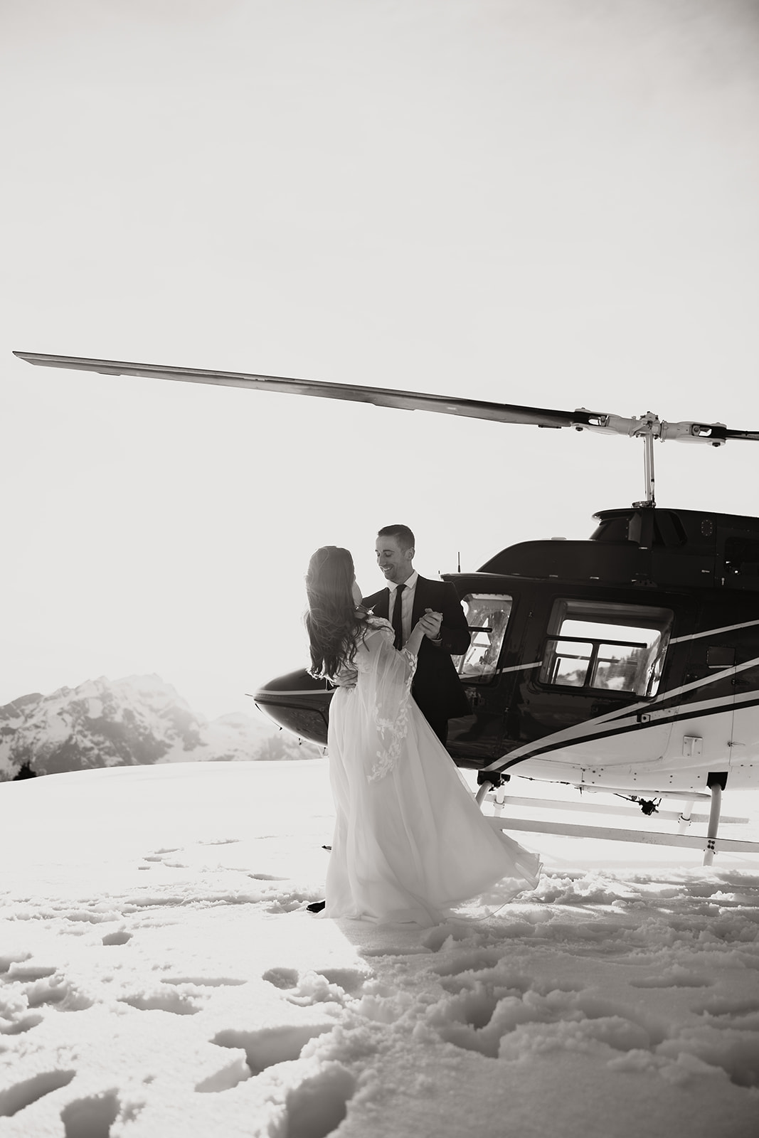 Black and white photo of Joyful couple holding hands and dancing by their helicopter, with the breathtaking mountain scenery surrounding them.