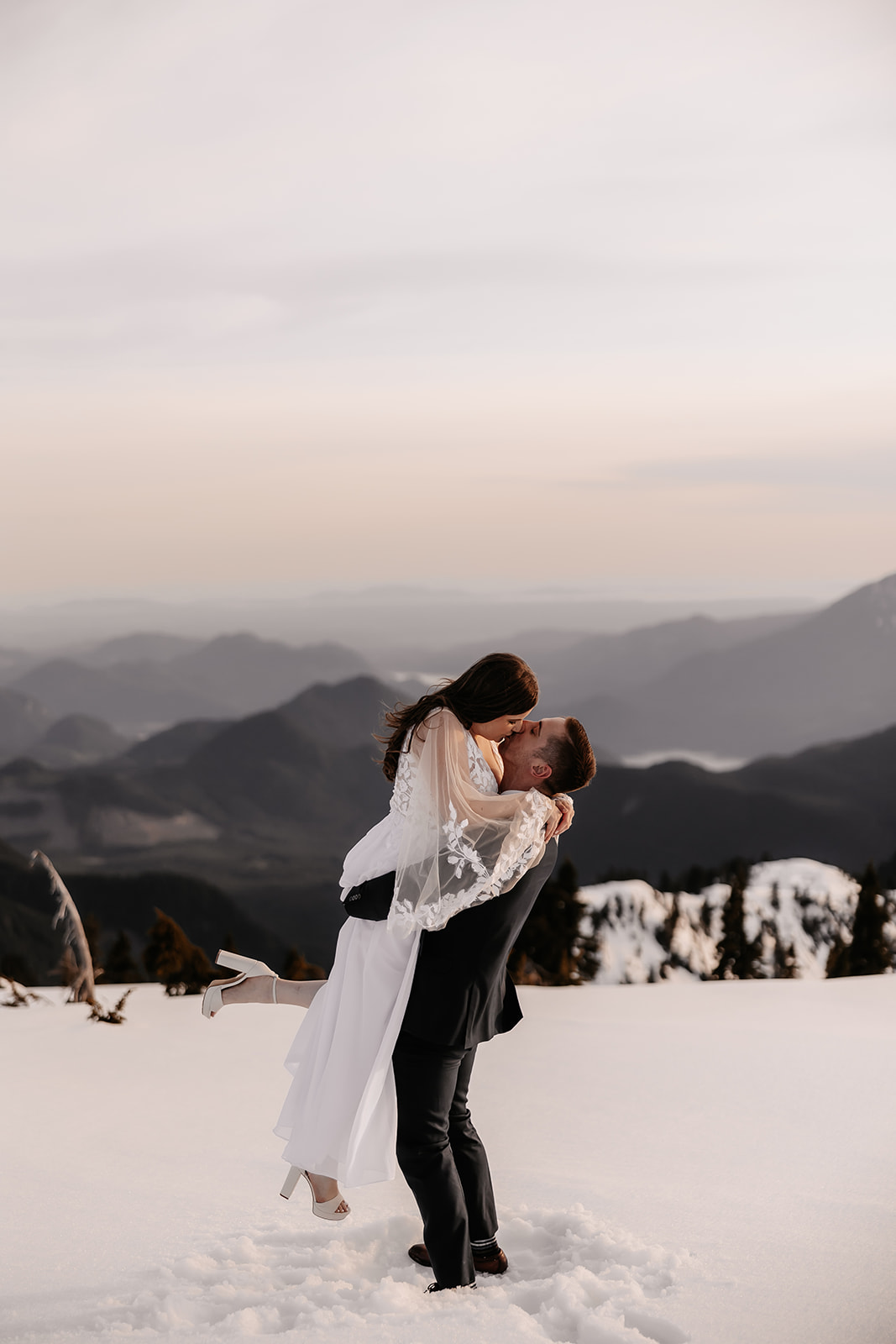 A joyful couple celebrating their helicopter elopement in the snowy mountains of British Columbia, sharing a loving kiss as they stand together against a breathtaking backdrop of peaks.