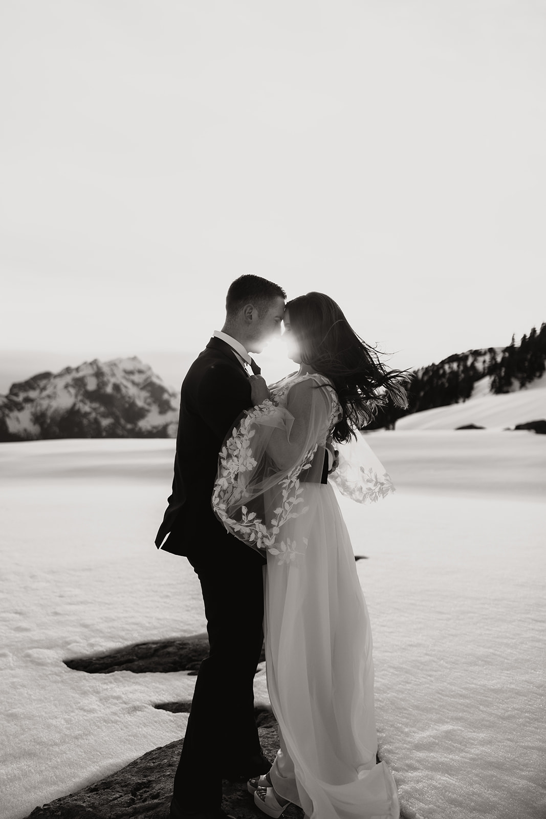 A black and white photo of a couple in formal attire sharing a tender moment, foreheads touching, surrounded by a snowy mountain landscape during their helicopter elopement.