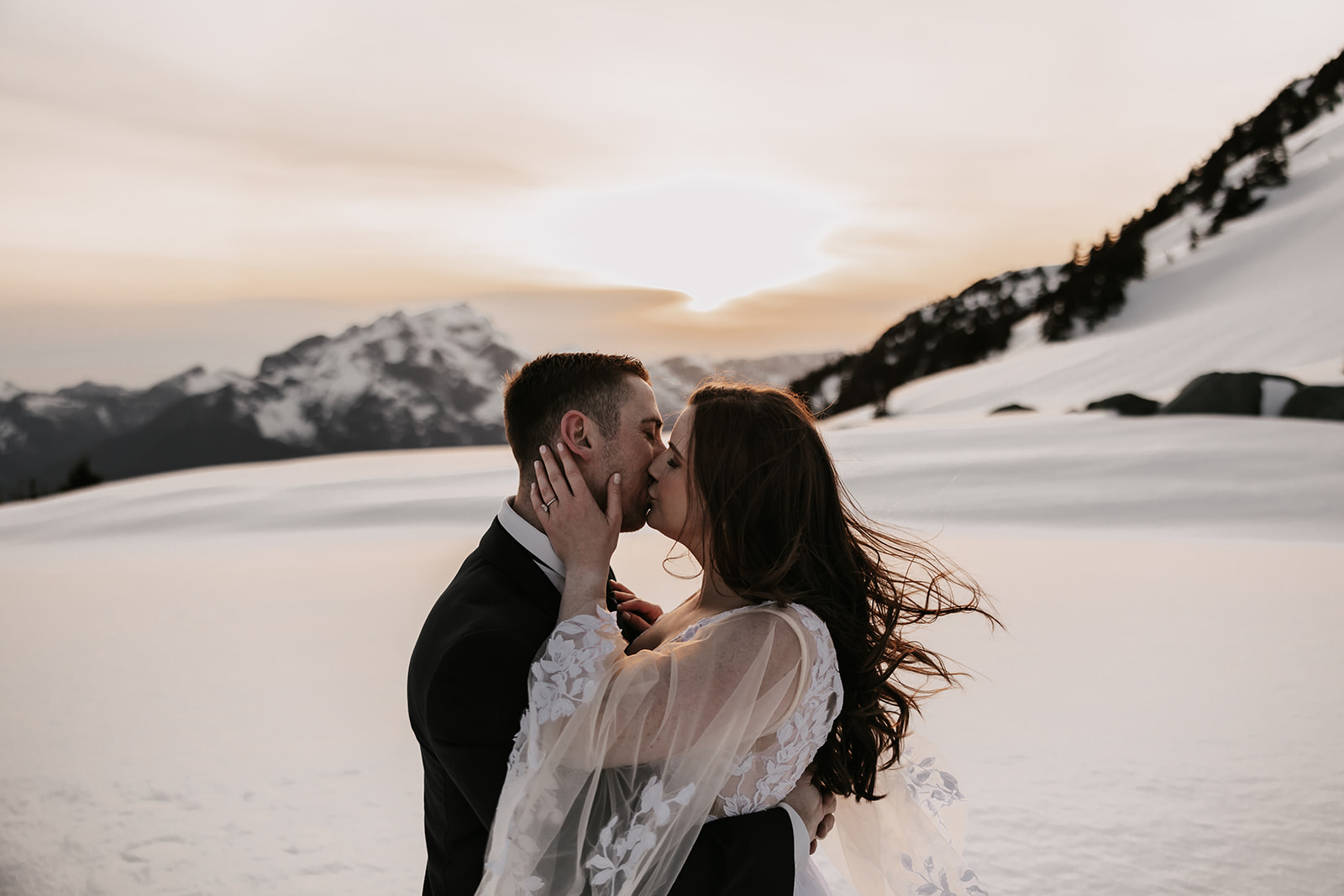 A couple kissing passionately in the snow, the sun setting behind the mountains, highlighting the romantic atmosphere of their helicopter elopement.