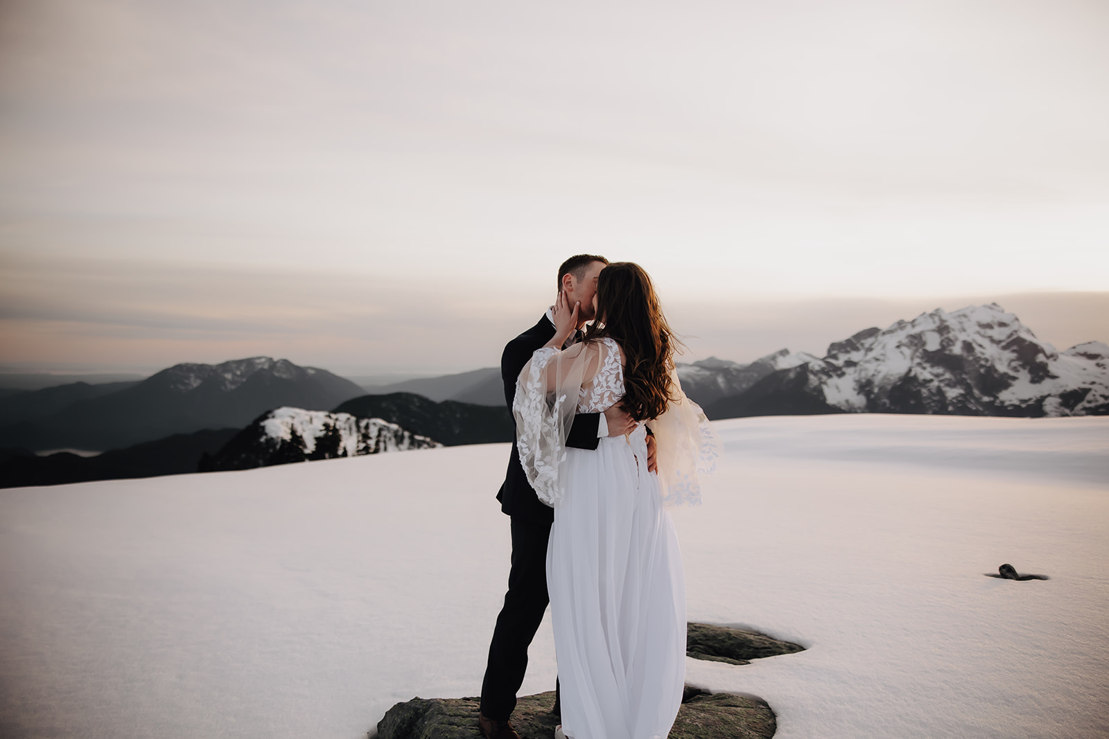 The couple embraces each other while standing on a snowy terrain, with majestic mountains in the background, celebrating their helicopter elopement adventure.