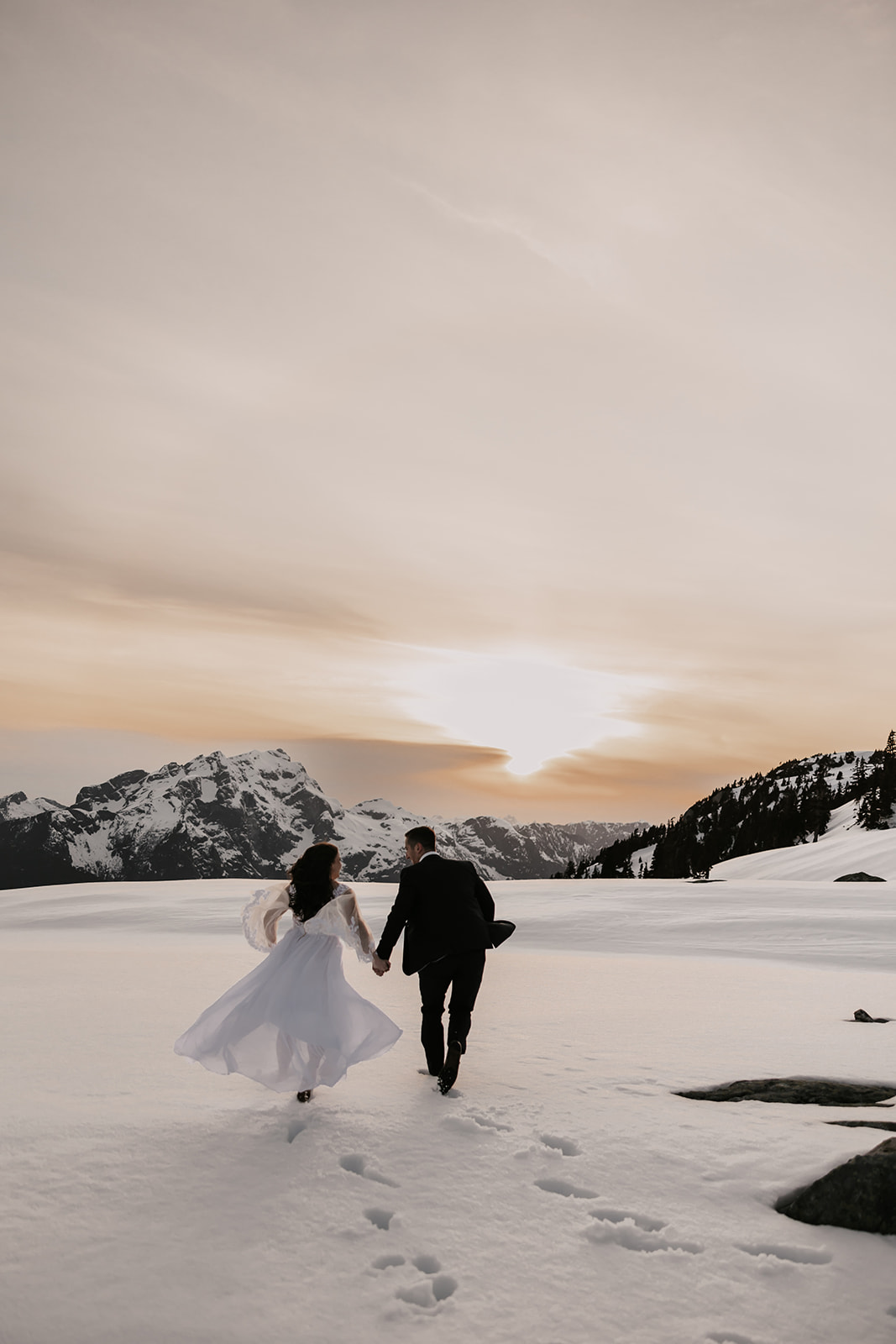 A couple walking hand-in-hand through a snowy field, with snow-covered mountains in the background and the sunset casting a warm glow, encapsulating the essence of their helicopter elopement.