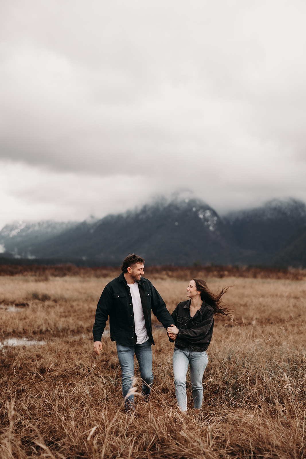 The couple stands close, sharing a laugh as they look at each other, surrounded by tall grasses. The moody clouds above add a dramatic touch to these fall engagement photos, showcasing the couple's joyful energy in a cozy outdoor setting.