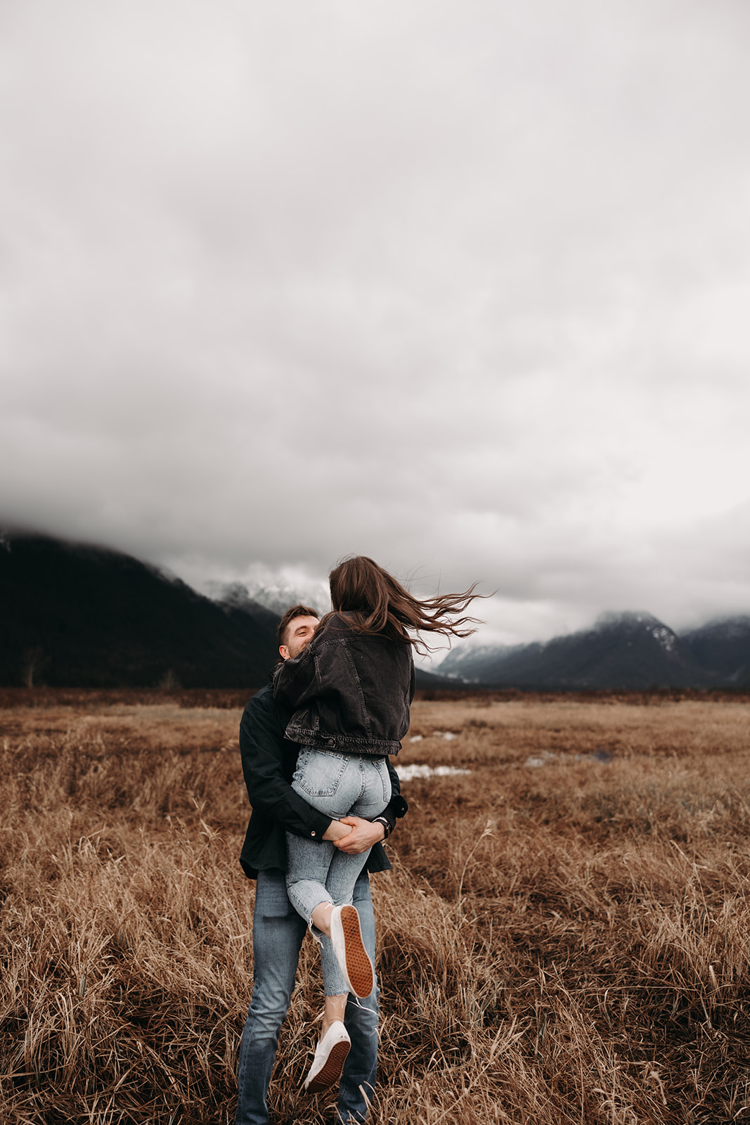 The couple shares a joyful embrace in this charming fall engagement photo, with the woman being playfully lifted by her partner. Their expressions radiate happiness against a backdrop of soft clouds and earthy tones, capturing the essence of love and fun during the autumn season.