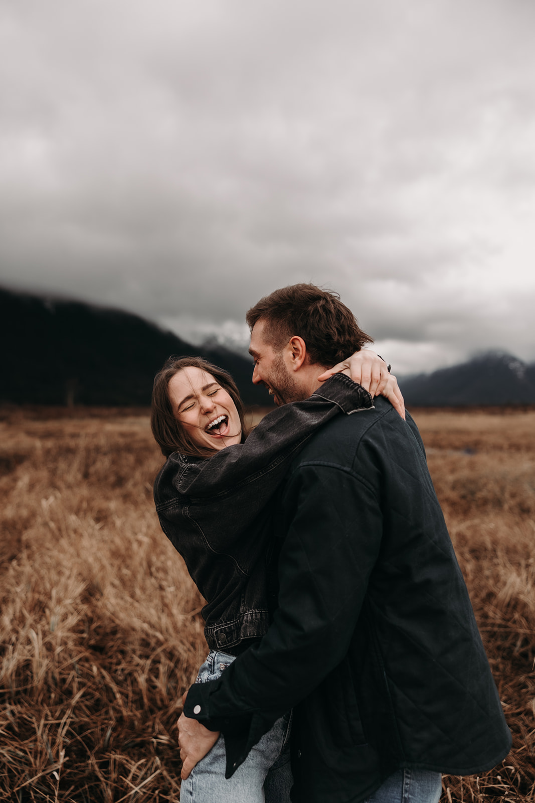 The couple embraces playfully, with the woman laughing and the man holding her close. This candid moment perfectly embodies the spirit of their fall engagement photos, set against a stunning backdrop of mountains and muted autumn colors.