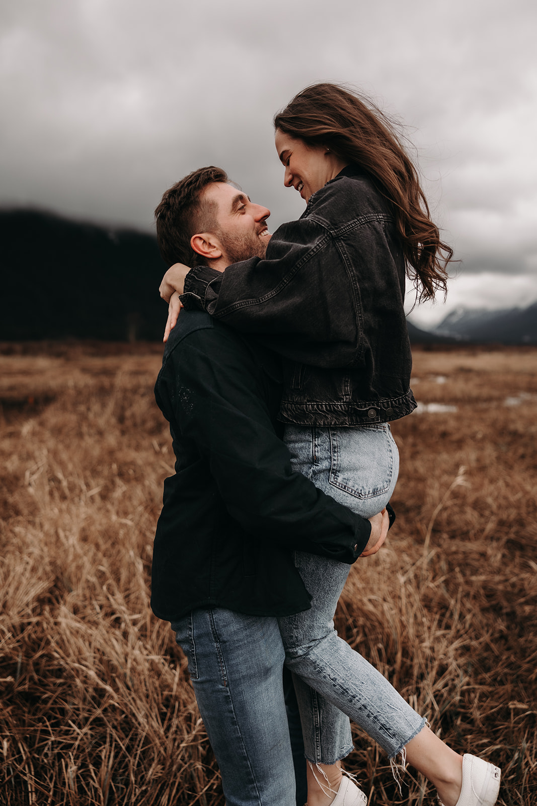 A happy couple enjoys a playful moment during their fall engagement photos as the man lifts the woman off the ground, showcasing their love and connection. The backdrop features a scenic landscape with muted autumn colors and mountains, highlighting the beauty of their intimate moment in the great outdoors.