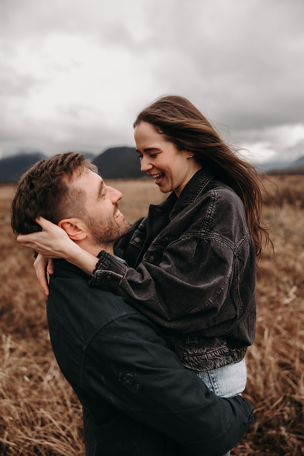  In this candid moment from their fall engagement photos, the couple smiles at each other while the man holds the woman securely in his arms. The surrounding nature adds depth to the image, with the muted colors of the grass and mountains creating a warm, inviting atmosphere that perfectly reflects their love story.