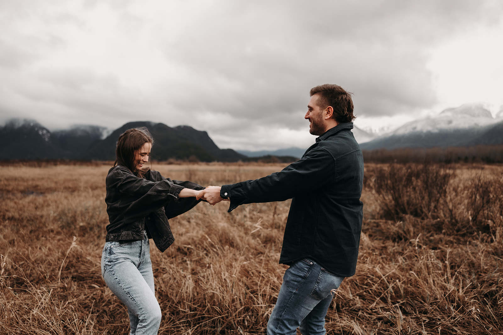 A candid capture of a couple playfully holding hands and spinning together in a field of tall grass. Their joyful expressions and the backdrop of majestic mountains create an enchanting setting for their fall engagement photos, showcasing the beautiful Pacific Northwest vibes.