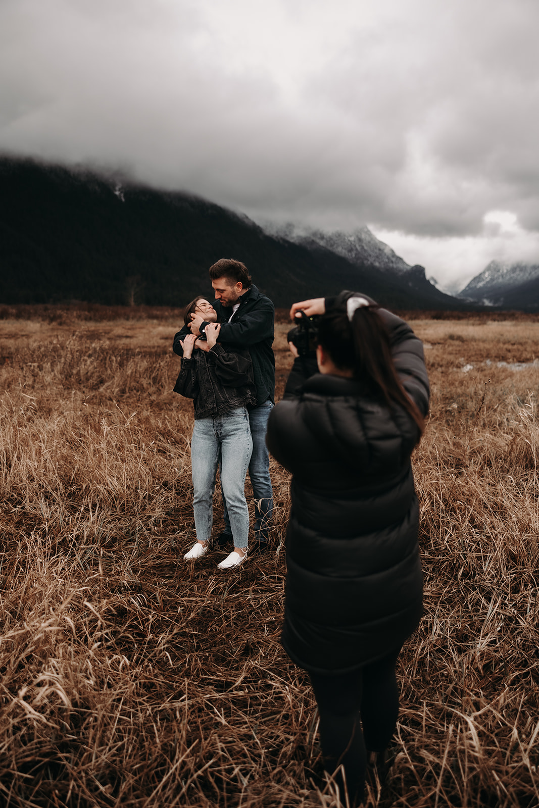 A candid shot of the couple in the foreground with the photographer's hand holding a camera in the background. They share a sweet moment, surrounded by the stunning landscape of Pitt Lake, showcasing the joyful spirit of their fall engagement photos 