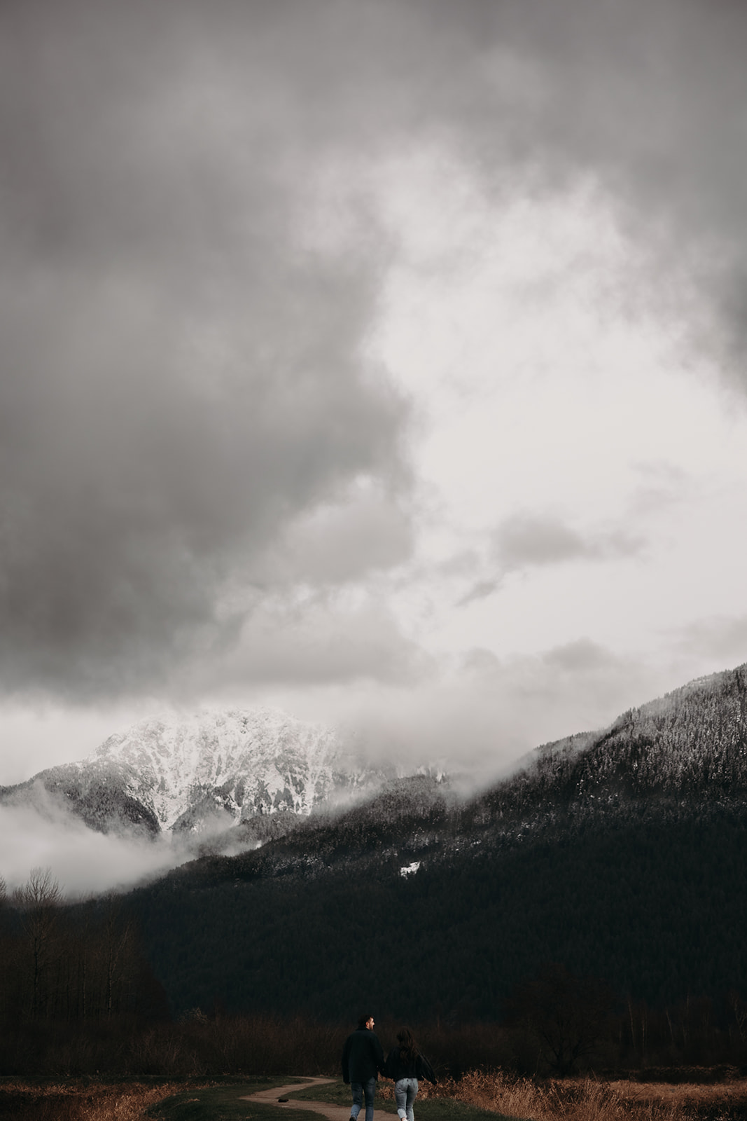 A couple walks hand in hand along a path surrounded by golden grass, with a majestic snow-capped mountain looming in the background. The overcast sky adds a cozy touch to this fall engagement photo, highlighting their intimate connection and the beauty of nature.