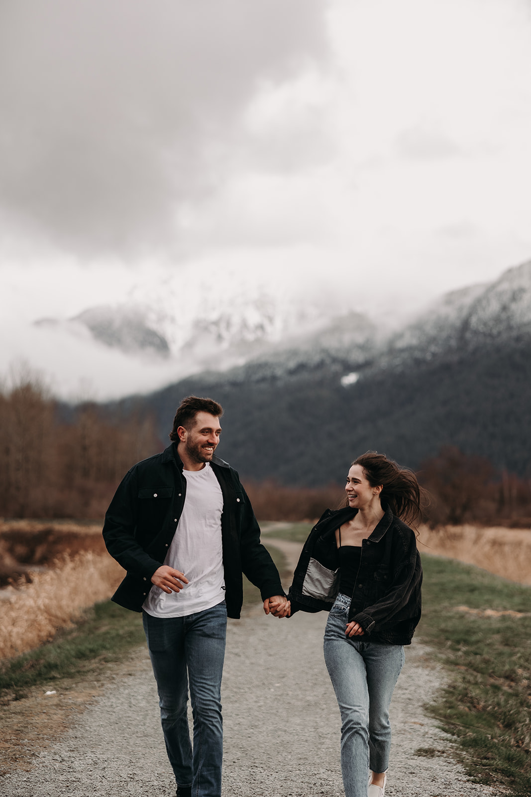  A couple joyfully walks hand-in-hand down a gravel path surrounded by golden grass and lush green hills. The man, wearing a black jacket and a white t-shirt, shares a genuine smile with the woman, who is dressed in a black jacket and light blue jeans. The cloudy sky and mountain backdrop set the mood for their fall engagement photos, showcasing the beauty of love in nature.