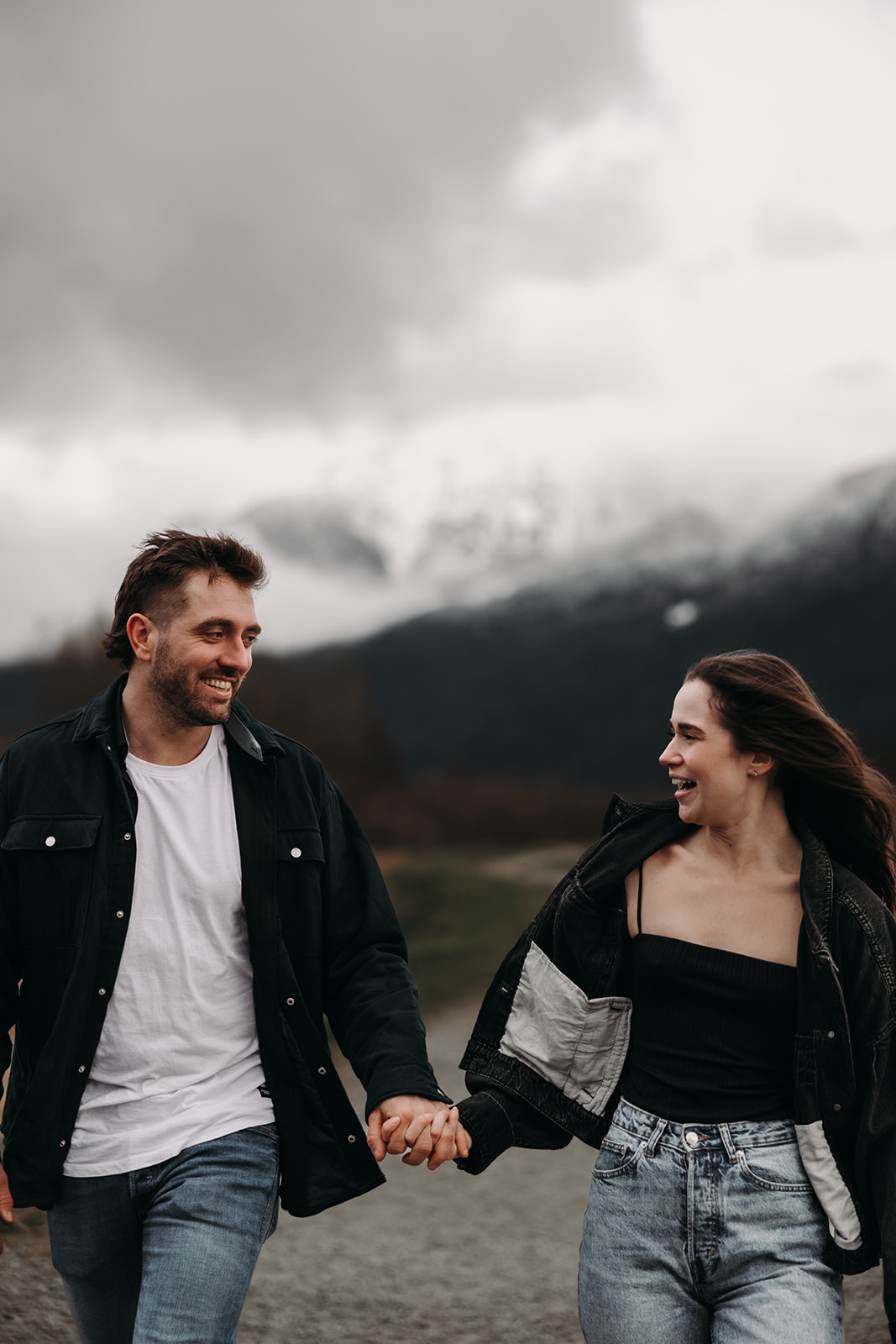 A couple walks hand-in-hand through a golden field with mountains in the background, their smiles brightening the cloudy day. These fall engagement photos beautifully capture their connection amidst the rich autumn hues of the landscape.