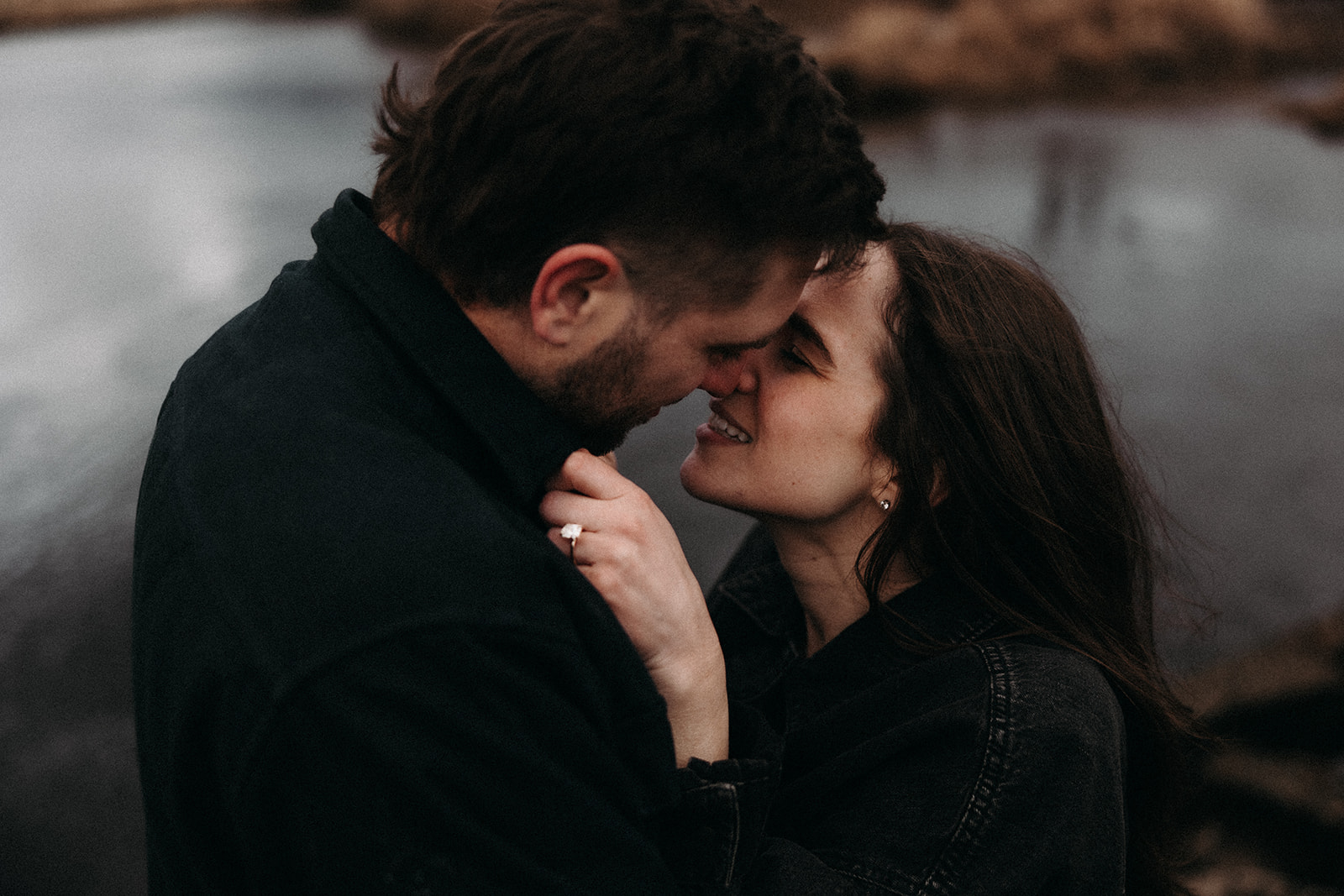 A close-up shot of a couple sharing a sweet moment, their foreheads touching as they smile at each other. The background features a blurred view of a serene lake and cloudy sky, perfect for fall engagement photos. The woman's engagement ring sparkles as she gently holds onto her partner's shirt.