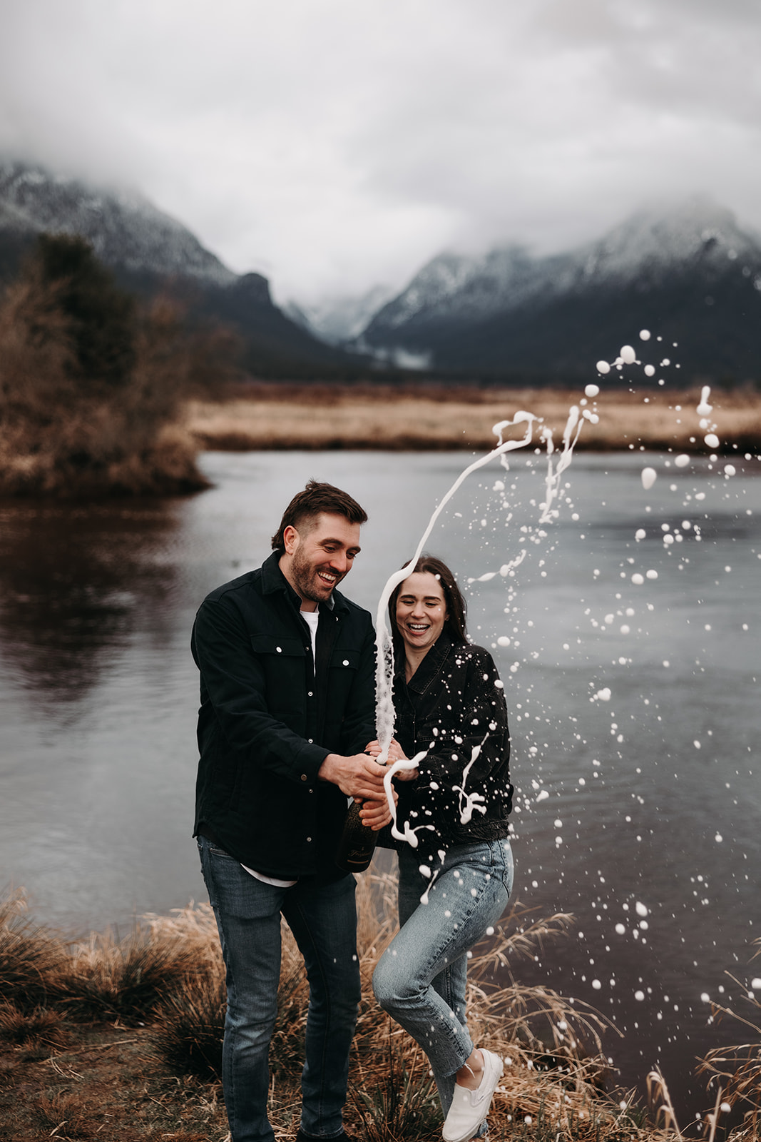 A joyful couple stands by the water, laughing as they spray sparkling cider into the air, creating a playful splash around them. This moment captures the fun and spontaneity of their fall engagement photos, set against the stunning backdrop of a cloudy sky and majestic mountains in the distance.